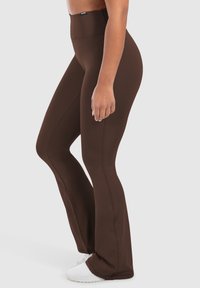 Donna che indossa leggings a vita alta, marrone scuro a zampa e sneakers bianche, in piedi di lato contro uno sfondo chiaro e uniforme.