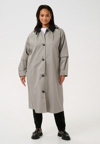 Grå knäppt trenchcoats med svart krage, med breda ärmar och två sidofickor, bärs över svarta byxor och kraftiga svarta skor.