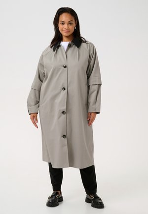 Grå knäppt trenchcoats med svart krage, med breda ärmar och två sidofickor, bärs över svarta byxor och kraftiga svarta skor.
