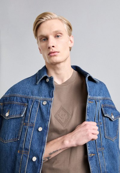 Chaqueta de mezclilla con botones plateados y dos bolsillos frontales, llevada sobre una camiseta marrón con un logo de diamante en relieve. Textura de tela suave.