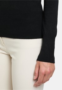 Schwarzer Strickpullover mit taillierter Passform, versehen mit einem gerippten Saum und Bündchen. Getragen über hellbeigen, enganliegenden Hosen.