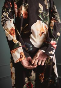 Šaty s květinovým vzorem v tmavé tkanině; obsahují velké, barevné květy. Dlouhé rukávy s červenými akcenty na manžetách; ruce složené za zády.