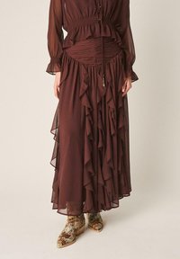 Femme portant une robe marron transparente à manches longues avec des volants et des détails froncés, associée à des bottines à motif serpent multicolores.