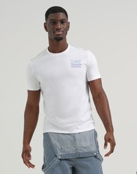 Uomo di colore che indossa una t-shirt bianca Lee Crayola e pantaloni a strisce blu con dettaglio in tessuto sovrapposto, in piedi contro uno sfondo chiaro e semplice.