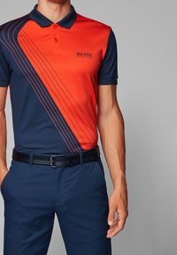 Polo tričko v námořnické modré a oranžové barvě s horizontálním pruhovaným designem. Krátké rukávy, límeček a logo na hrudi. Nosit s černým opaskem.