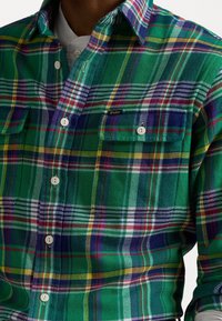 Camicia a quadri verde con strisce bianche, viola e gialle, dotata di due tasche frontali e chiusura con bottoni bianchi.