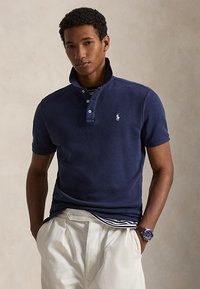 Vyras dėvi tamsiai mėlyną trumpomis rankovėmis polo marškinėlį su pakelta apykakle, smėlio spalvos kelnes, po jais dryžuotą marškinių sluoksnį ir mėlyną riešo laikrodį, stovintis priešais paprastą foną.