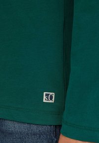 Grünes Langarmshirt aus weichem Stoff mit einem kleinen weißen Etikett in dunkler Schrift, vor blauen Jeans präsentiert.