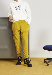 Felpa bianca con logo, pantaloni jogger gialli e sneaker nere. I pantaloni jogger hanno un cintura elastica e tasche laterali, le sneaker presentano accenti neri e gialli.