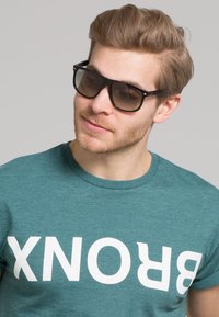 Tricou din bumbac teal cu imprimeu alb îndrăzneț "BRONX", asortat cu ochelari de soare închiși la culoare. Design cu mâneci scurte și guler rotund. Aspect lejer.