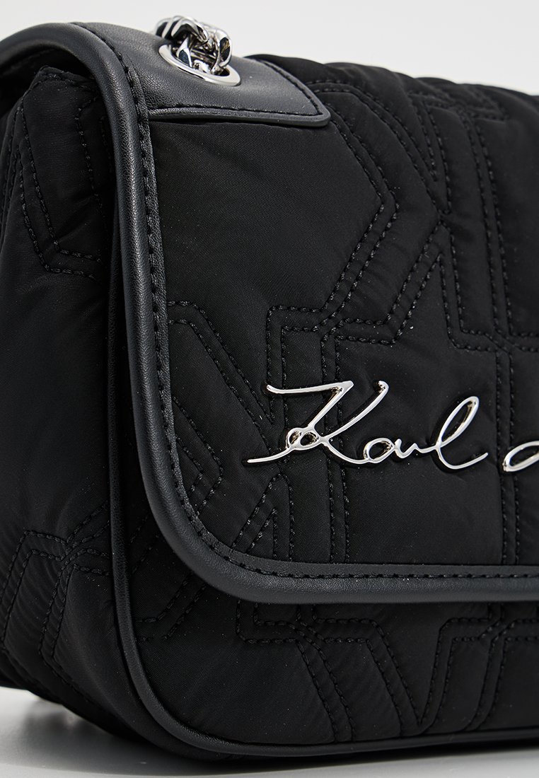 Sac à main noir en cuir matelassé avec une couture géométrique et un logo en métal argenté sur le rabat avant, avec des finitions en cuir et du matériel en métal.