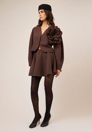 Femme portant un tailleur jupe marron avec un grand détail floral sur l'épaule, collants noirs, talons hauts noirs et un béret noir, debout devant un fond uni.