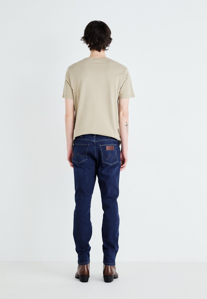 T-shirt in cotone beige con maniche corte; abbinata a jeans blu scuro con un'etichetta in pelle e tasche posteriori con dettagli cuciti.