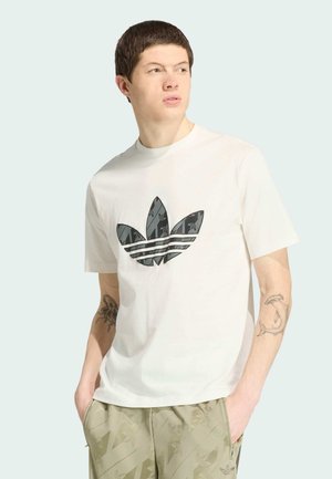 Mladić s kratkom tamnom kosom, obučen u bijelu Adidas majicu s crnim logom, bež uzorkane hlače i tetovaže na oba podlaktica, gleda lijevo.