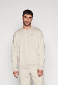 Sudadera beige clara con cuello redondo, mangas largas y puños acanalados. Presenta un pequeño logo bordado en el pecho. Textura suave.