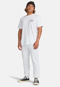 Camiseta blanca de manga corta con un logo circular multicolor, combinada con pantalones de chándal grises y zapatillas negras con cordones blancos.