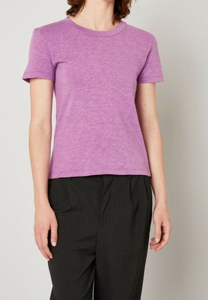 Mujer vestida con una camiseta de manga corta lavanda con cuello redondo y pantalones plisados negros frente a un fondo claro y liso.