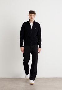 Svart zip-up cardigan med en strukturerad yta, bärs över en vit t-shirt, kombinerad med lösa svarta byxor och vita sneakers.