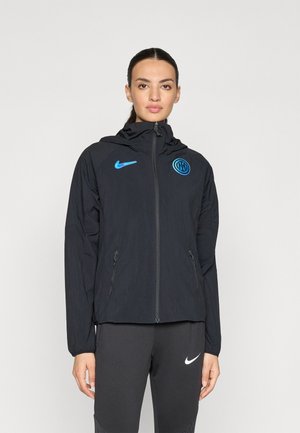 Juoda Nike striukė su aukštu apykakle, pilnu užtrauktuku ir mėlynu logotipu. Yra dvi šoninės kišenės ir lengvas audinys su tekstūruota apdaila.