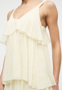 Αμάνικο κρεμ φόρεμα με στρωματοποιημένα βολάν στο πάνω μέρος, φορεμένο από άτομο με ακάλυπτους βραχίονες και ώμους.