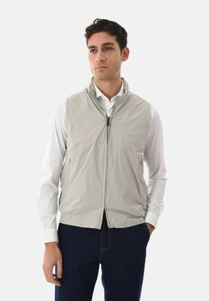 Uomo che indossa un gilet grigio chiaro senza maniche con cerniera sopra una camicia bianca a maniche lunghe e pantaloni scuri, in piedi con una mano in tasca.