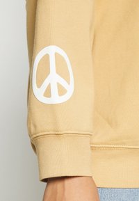 Sudadera beige con una textura suave, que presenta un gran símbolo de la paz blanco en la manga izquierda. El puño tiene un acabado acanalado.
