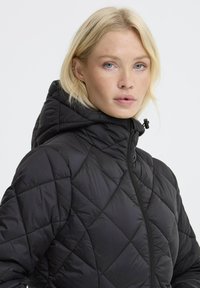 Femme blonde aux yeux bleus portant une veste à capuche matelassée noire zippée contre un fond clair.
