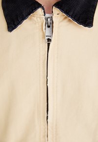 Beżowa kurtka z zamkiem błyskawicznym z przodu. Posiada granatowy kołnierz z sztruksu i teksturowaną tkaninę. Charakterystyczny metalowy zamek błyskawiczny z logo "Weekday".