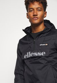 Schwarze leichte Pulloverjacke mit einem Reißverschlusskragen, elastischen Bündchen und einem auffälligen grauen "ellesse"-Logo auf der Brust. Texturiertes Material.