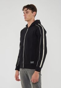 Felpa nera con zip, con strisce bianche sulle maniche, dotata di tasca frontale e patch con logo in basso. Tessuto morbido.