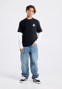 Mustat lyhythihaiset t-paidat, jotka on kerroksittain valkoisen pitkähihaisen paidan päällä, yhdistetty vaaleansinisiin farkkuihin sekä mustiin ja valkoisiin lenkkareihin. Etutaskun logo-yksityiskohta.