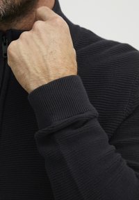 Sötét bordás kötött pulóver, magas gallérral és szűk mandzsettákkal. A textúra sima, finom függőleges vonalakkal az anyagon.