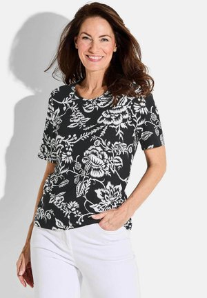 Haut à manches courtes noir à motifs floraux avec des dessins blancs, présentant un col rond et une texture subtile, associé à un pantalon blanc.