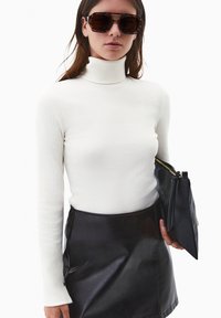 Biała żebrowana bluzka z golfem, czarna skórzana mini spódniczka z bocznymi guzikami, trzymająca czarną kopertówkę. Dodatki to przesadzone okulary w panterkę.