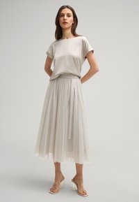 Helles beiges Satinoberteil mit aufgerollten Ärmeln; passender plissierter Tüllrock mit einer taillierten Taille. Klare Absatzsandalen runden den Look ab.