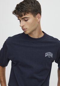 Marineblå bomulds T-shirt med korte ærmer, rund hals, med et hvidt "CASUAL" logo trykt på venstre brystområde. Glat tekstur.