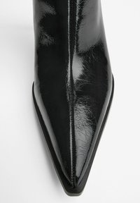 Gheata de gleznă din piele lacuită neagră, cu vârf ascuțit, textură netedă și cute subtile. Prezintă un design minimalist și o talpă subțire.