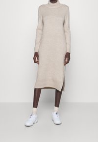 Abito a maglia beige con un collo alto e maniche lunghe, con uno spacco laterale. Indossato con sneaker bianche e calze crema.