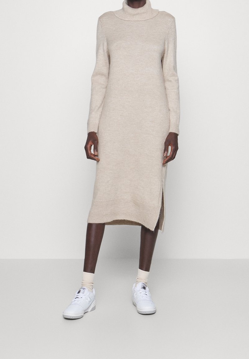 Abito a maglia beige con un collo alto e maniche lunghe, con uno spacco laterale. Indossato con sneaker bianche e calze crema.