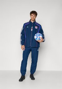 Marine idrettssett med hvite striper og logoer, bestående av en zip-up jakke og bukser med elastisk liv, kombinert med en fotball.