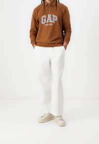 Pullover in pile marrone con logo "GAP" e zip a metà, abbinato a pantaloni larghi in lino bianchi e sneakers beige.