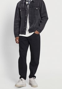 Hombre con chaqueta de mezclilla negra, camisa blanca, pantalones negros y zapatillas blancas, con una mano en el bolsillo, frente a un fondo claro y liso.