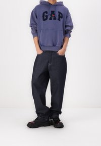 Person som bär en lila GAP-hoodie med detalj av en röd ros, mörka löst sittande jeans och svarta klumpiga skor, står med händerna i fickorna.