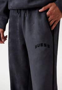 Sötét szürke bolyhos jogger nadrág, puha tapintással. Zsebekkel és fekete "GUESS" logóval a combon, kontrasztos oldalsó díszítéssel.