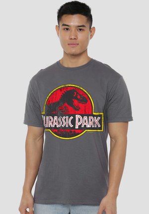 Young man wearing gray Jurassic Park T-shirt with red and yellow dinosaur logo on a plain light background.