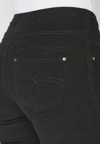 Pantalones de pana negros con una cintura alta, bolsillos traseros con remaches plateados y un detalle de costura distintivo en el bolsillo.