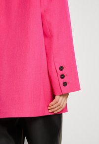 Ljusrosa jacka med en slät yta, med skräddarsydd passform och fyra svarta knappar på ärmen. Svarta, glansiga byxor synliga.