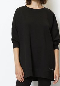 Sudadera negra extragrande con cuello redondo, hombros caídos y mangas largas. Presenta una etiqueta de logo cerca del dobladillo. Tejido liso y texturizado.