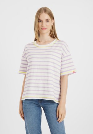 Blondynka w pastelowo fioletowo-białej koszulce w paski z krótkim rękawem i niebieskich dżinsach, zwrócona przodem na jednolitym białym tle.