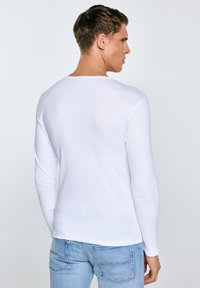 Vīrietis ar īsiem brūniem matiem, valkājot piegulošu baltu garā piedurknes kreklu un gaismazilas džinsas, skatās prom pret vienkāršu fonu.